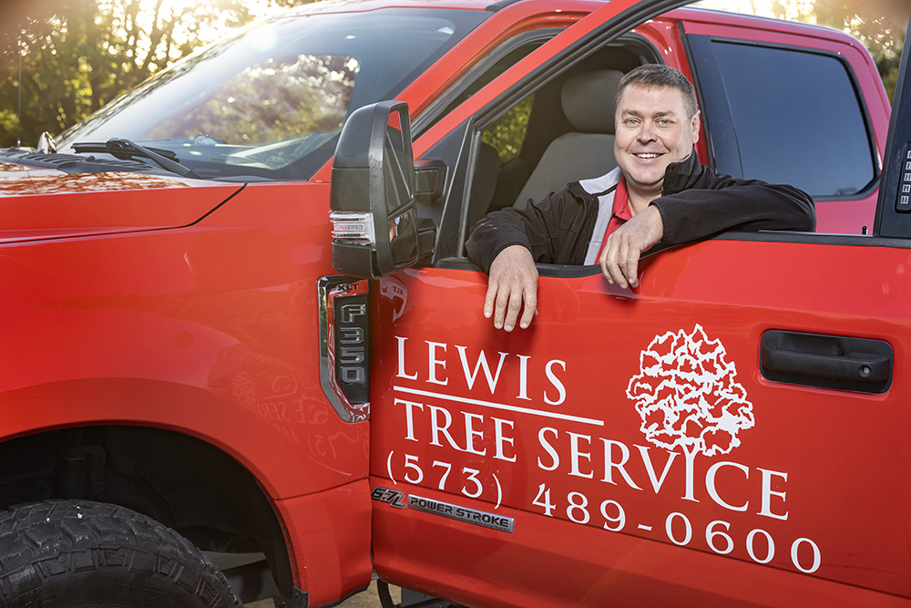 Matt Lewis rests his arms on the open window of his driver side door. The company logo for Lewis Tree Service is prominently displayed on the door of the truck. 